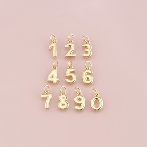 May include: A collection of gold-colored number charms, each with a small loop at the top for attaching to a chain or bracelet. The numbers 1 through 0 are displayed against a soft pink background. These charms are ideal for personalized jewelry.