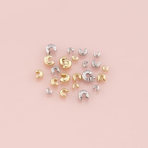 May include: A collection of small, C-shaped jewelry findings in gold and silver tones. These findings are scattered on a light pink surface, showcasing their curved design and metallic sheen. Ideal for jewelry making and crafting.