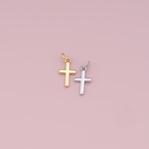 Puede incluir: Dos pequeños dijes de cruz, uno dorado y otro plateado, sobre un fondo rosa.