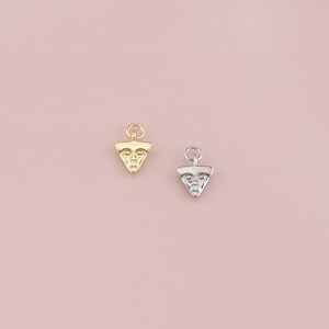 May include: Two small, triangular pendants with a face design. One is gold-colored, the other is silver-colored. Both have a small loop at the top for attaching to a chain or bracelet. The pendants are displayed against a soft pink background.