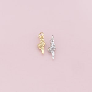 May include: Two seashell charms, one gold-colored and one silver-colored, are displayed against a soft pink background. The charms are detailed with textured surfaces, showcasing the intricate patterns of a seashell. These pendants are suitable for jewelry making.