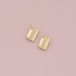 Puede incluir: Dos charms de cinta de casete de tono dorado con acentos de diamantes de imitación transparentes.