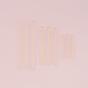 Peut inclure: Plusieurs épingles à tête dorées de différentes longueurs, disposées sur un fond rose clair. Les épingles ont une tige droite et fine avec une petite tête plate. Ces épingles sont probablement utilisées pour la fabrication de bijoux ou l'artisanat.
