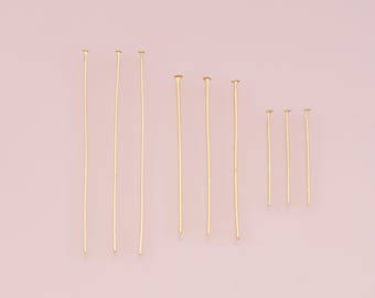May include: A collection of gold-colored head pins of varying lengths, arranged on a soft pink background. The pins have a straight, slender shaft with a small, flat head. These pins are likely used for jewelry making or crafting.