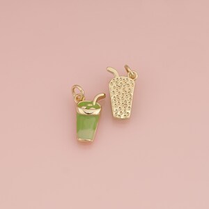 May include: Two gold-toned metal charms shaped like cups with straws. One charm features a smooth light green enamel finish, while the other has a textured gold surface with small raised dots, both including a small jump ring for attachment.
