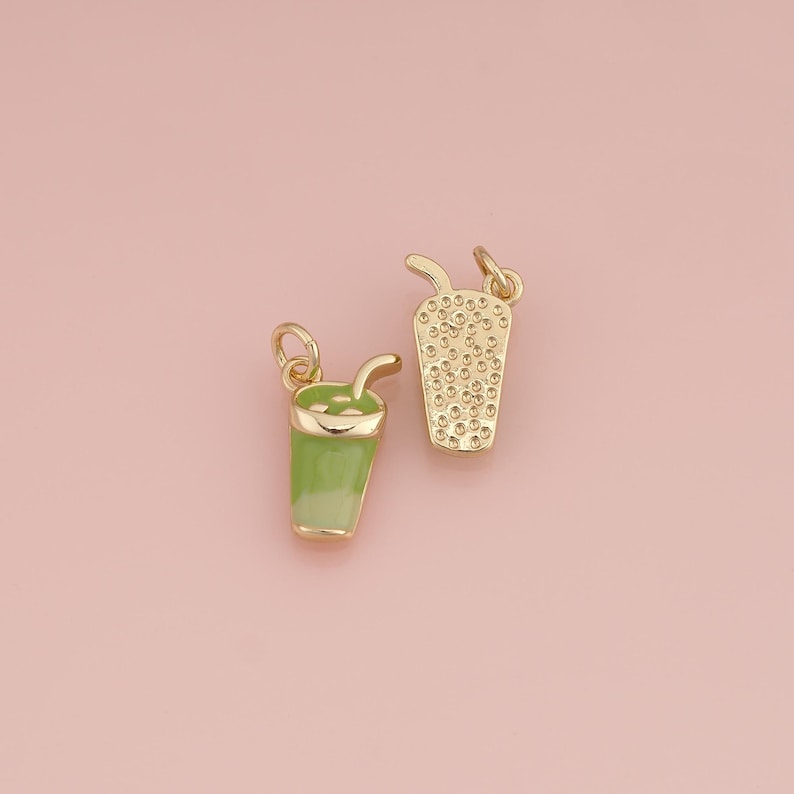 May include: Two gold-toned metal charms shaped like cups with straws. One charm features a smooth light green enamel finish, while the other has a textured gold surface with small raised dots, both including a small jump ring for attachment.