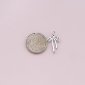 May include: Silver charm in the shape of a bow with a single rhinestone in the center. The charm is about the size of a US quarter.
