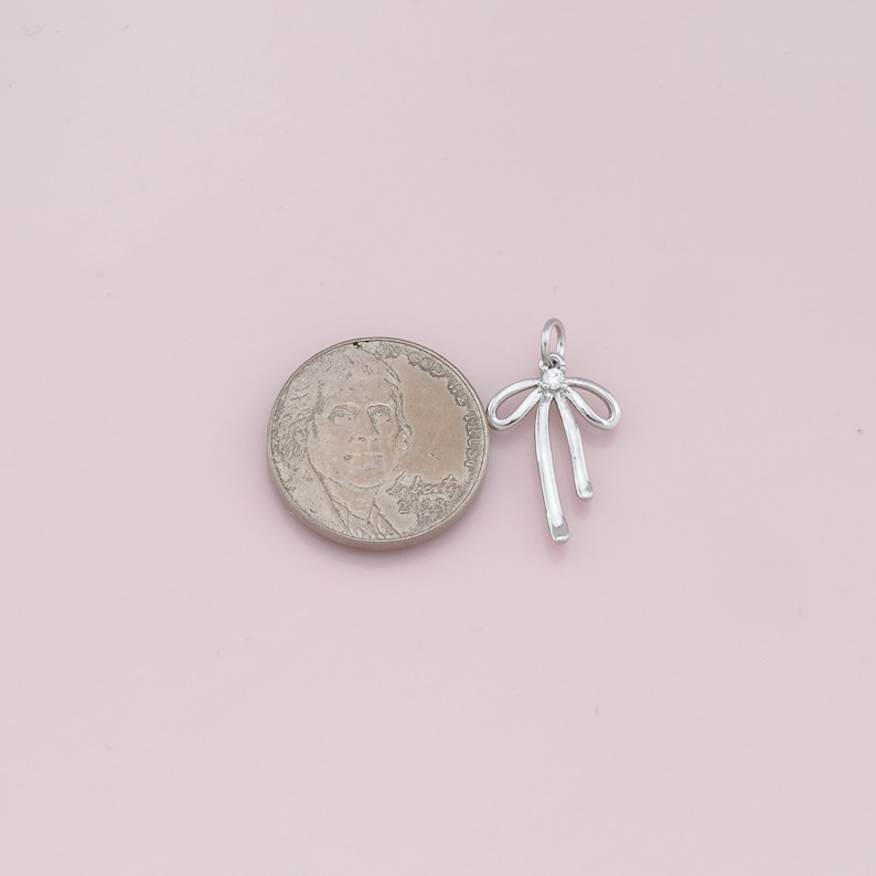 May include: Silver charm in the shape of a bow with a single rhinestone in the center. The charm is about the size of a US quarter.