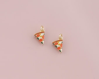 May include: Two gold-coloured pizza slice charms with red, white, green, and yellow enamel detailing. The charms are on a light pink background. Each charm has a small loop at the top for attaching to a chain or bracelet.