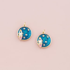 May include: Two gold-toned charms with a blue enamel background depicting a white rocket ship flying through a starry night sky. The charms feature a crescent moon and stars.
