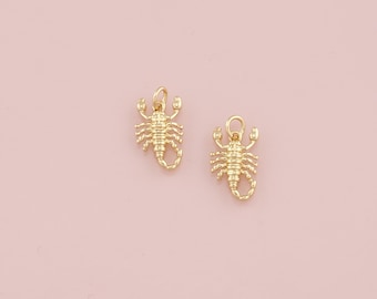 Puede incluir: Dos colgantes dorados con forma de escorpión. Cada colgante presenta un diseño detallado de escorpión con un lazo en la parte superior para sujetarlo a un collar o pulsera. Los colgantes están sobre un fondo rosa suave.