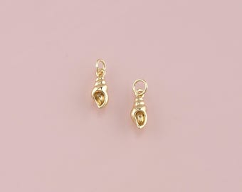 Puede incluir: Dos pequeños colgantes dorados con forma de concha, con un lazo para colgar. Los colgantes presentan detalles de estrías y un interior hueco, sobre un fondo rosa claro. Adecuado para la creación de joyas.