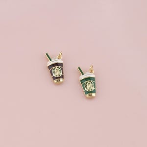 以下が含まれることがあります： 緑と茶色のエナメルディテールが施された、2つの金色のコーヒーカップチャーム。各チャームにはストローとロゴが付いています。チャームはソフトピンクの背景に配置されています。