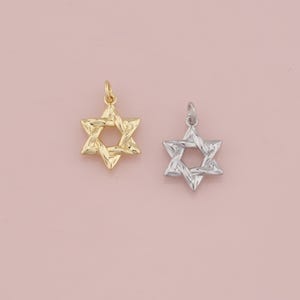 May include: Two Star of David pendants. One is gold-colored, the other is silver-colored. Both pendants have a textured surface and a loop at the top for attaching to a chain. The background is a solid light pink.