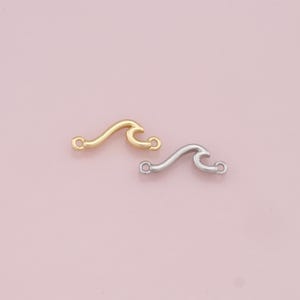 May include: Two wave-shaped metal charms with jump rings. The charm on the left is gold and the charm on the right is silver.