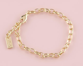 May include: A gold-colored chain bracelet with an oval link design. The bracelet has a lobster clasp closure and an adjustable chain. A small rectangular tag with text is attached to the chain. The bracelet is displayed on a pink background.
