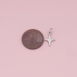 May include: A silver charm in the shape of a four-pointed star with clear cubic zirconia stones. The charm is shown next to a US penny for size comparison.