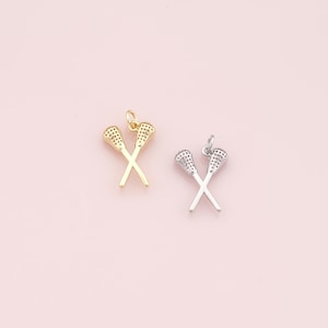 May include: Two lacrosse stick charms, one gold-colored and one silver-colored, are displayed against a soft pink background. The charms are crossed, with detailed netting on the stick heads. These pendants are ideal for jewelry making.