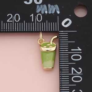 May include: A gold-toned metal charm shaped like a cup with a straw, featuring light green enamel detailing. The charm is positioned next to a black ruler, showing a height of approximately 0.5 inches from the top of the straw to the base.