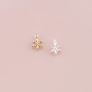 Op de afbeelding: Twee sneeuwvlokbedels, één goudkleurig en één zilverkleurig, elk versierd met kleine, heldere kristallen. De bedels worden gepresenteerd tegen een zachtroze achtergrond, die hun delicate ontwerp en sprankelende details benadrukt. Ideaal voor het maken van sieraden.
