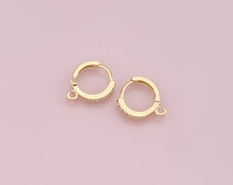 Puede incluir: Dos pendientes de aro dorados con pequeñas piedras preciosas transparentes engastadas a lo largo de la mitad delantera de los aros. Cada pendiente tiene un pequeño lazo en la parte inferior para sujetar abalorios. Fondo rosa claro.