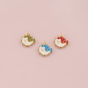 Op de afbeelding: Drie kleurrijke kogelvisbedels. Elke bedel heeft een goudkleurige metalen omtrek met een witte body, stekelige details en een kleine lus om aan sieraden te bevestigen. Eén is groen, één is blauw en één is rood.