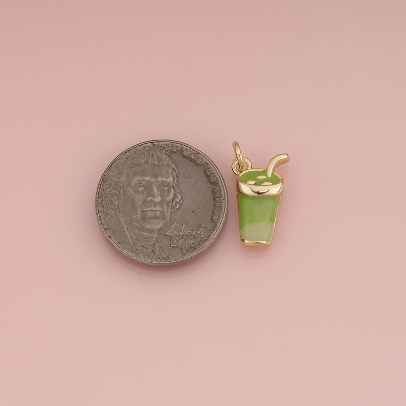 May include: A gold-toned metal charm shaped like a cup with a straw, featuring light green enamel filling. The charm is positioned next to a United States nickel coin for scale, showing the relative size of the small beverage-themed jewelry piece.
