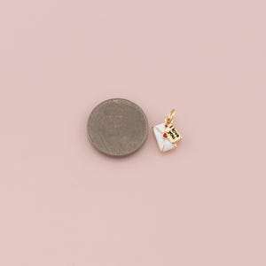 May include: A gold-colored charm in the shape of a white envelope with a red heart and the words "I love you" on a small tag. The charm is next to a coin on a pink background.