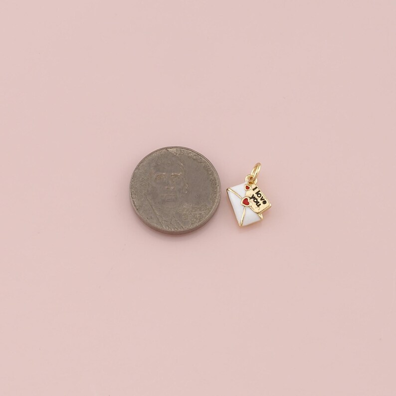 May include: A gold-colored charm in the shape of a white envelope with a red heart and the words "I love you" on a small tag. The charm is next to a coin on a pink background.