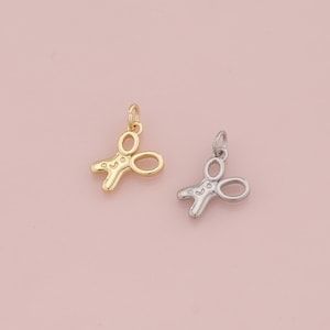 May include: Two small scissor charms, one gold-colored and one silver-colored, are displayed against a soft pink background. The scissors have a simple, open design with a loop at the top for hanging. These charms are suitable for jewelry making.