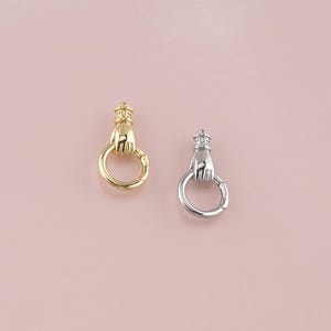 May include: Two metal clasps, one gold-toned and one silver-toned, each featuring a hand holding a ring. The hand is adorned with a crown. The clasps are likely for jewelry or accessories.