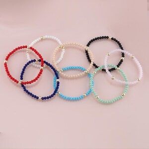 May include: A collection of beaded bracelets in various colors, including red, white, blue, black, beige, and turquoise. Each bracelet features small, round beads and gold-colored accents. The bracelets are displayed on a light pink surface.