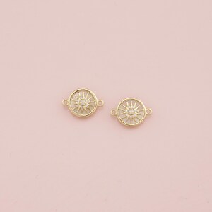May include: Two gold-colored sunburst charms with a central crystal and a ring of smaller crystals. Each charm has two small loops for attaching to jewelry. The charms are set against a soft pink background.