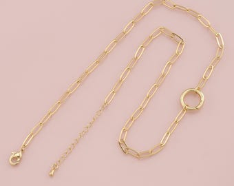 May include: A gold-colored necklace with an oval link chain design. The necklace features a large circular pendant and a lobster clasp closure. The chain is adjustable. The jewelry is displayed on a light pink background.
