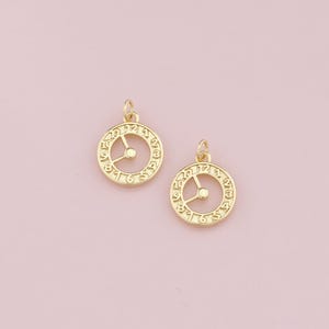 Puede incluir: Dos dijes de cara de reloj de tono dorado con marcas de números romanos. Los dijes tienen un círculo pequeño en el centro y una línea que se extiende desde el centro hasta el borde del círculo.