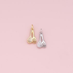 May include: Two charm pendants in the shape of a spoon and fork, both with a clear crystal design. The spoon and fork are joined together at the top. One pendant is gold and the other is silver.