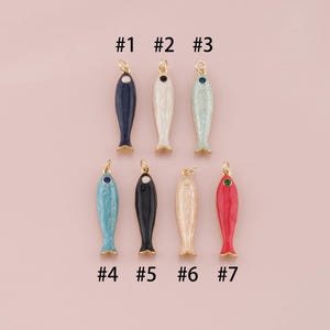 May include: Seven colorful fish-shaped charms with gold-colored accents. The charms are in various colors, including navy, white, light blue, black, cream, and red. Each charm has a small loop at the top for attaching to jewelry or other crafts. The image also includes the numbers #1 through #7.