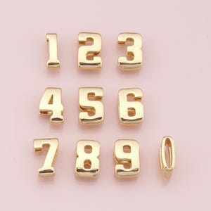 May include: A collection of gold-colored metal number charms, including the numbers 1 through 9 and a zero charm. The charms are in a bold, block style and are displayed against a soft pink background.
