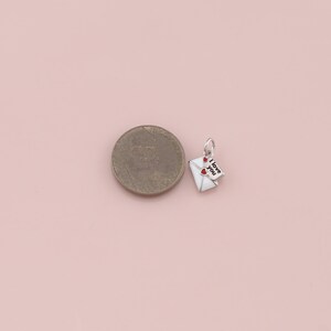 May include: A small, silver-toned charm in the shape of a white envelope with two red hearts and a small tag that reads "I love you". The charm is next to a coin on a light pink surface.