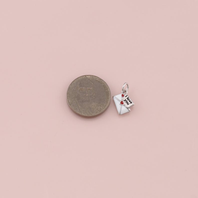 May include: A small, silver-toned charm in the shape of a white envelope with two red hearts and a small tag that reads "I love you". The charm is next to a coin on a light pink surface.