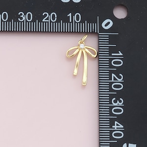 May include: A gold-toned bow charm with a clear rhinestone in the center. The charm is about 1 inch tall and is shown next to a ruler.