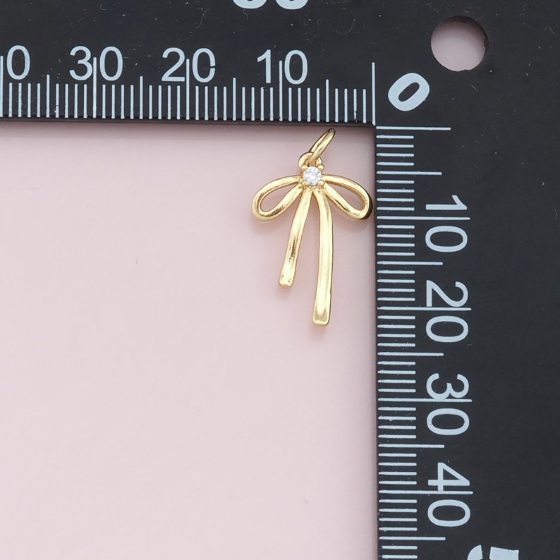 May include: A gold-toned bow charm with a clear rhinestone in the center. The charm is about 1 inch tall and is shown next to a ruler.
