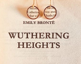 May include: A pair of gold-toned circular drop earrings featuring segments of printed book text from Wuthering Heights by Emily Brontë, with the book title and author name clearly visible in the background behind the jewelry pieces.
