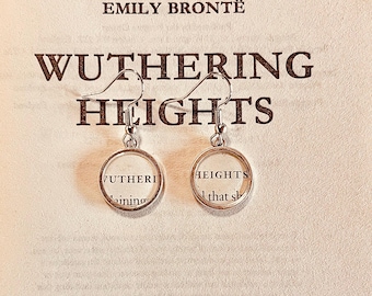 May include: A pair of silver-toned drop earrings featuring circular frames that enclose small paper cutouts with text from the book Wuthering Heights by Emily Brontë. The earrings hang from standard silver-toned metal hooks.