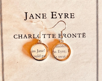 May include: A pair of silver-toned circular drop earrings featuring small segments of printed book text from Jane Eyre by Charlotte Brontë. The earrings hang from simple silver hooks and are displayed against the cover of the book.