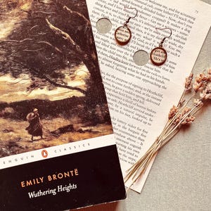 May include: A pair of round bronze-toned earrings featuring miniature text from the novel Wuthering Heights by Emily Brontë, displayed alongside a copy of the book and a small bundle of dried lavender stems on a neutral textured surface.