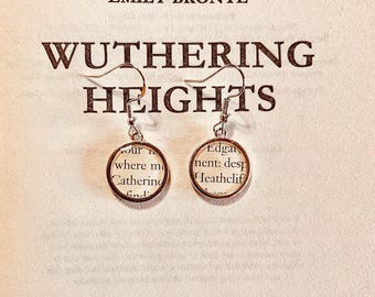 May include: A pair of silver-toned drop earrings featuring circular frames that enclose small, circular cutouts of printed book text. The earrings hang from standard silver-toned metal hooks and are displayed over the title Wuthering Heights.