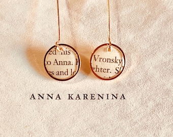 May include: A pair of rose gold drop earrings featuring circular frames that enclose small segments of printed book pages. The left earring shows text including 'ed his' and 'to Anna', while the right earring displays 'Vronsky' and 'hter'. The text 'ANNA KARENINA' is printed below.