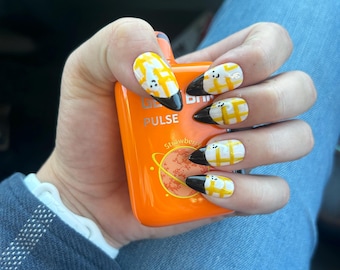 Peut inclure: Gros plan sur une main avec un art des ongles à carreaux noirs et jaunes. Les ongles sont longs et en forme d'amande. Le motif à carreaux est de couleur jaune clair et noir. Les ongles sont également décorés de petits points blancs. La main tient une petite bouteille orange avec le mot "PULSE" et le texte "Strawberry" dessus.