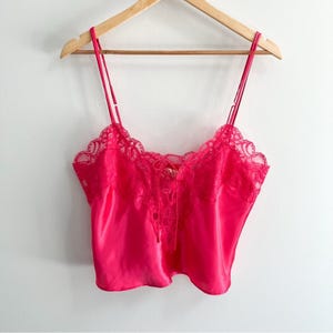 May include: A bright pink camisole with thin straps and a lace trim along the neckline. The camisole has a satin-like sheen and a tie detail in the center. It is hanging on a wooden hanger.